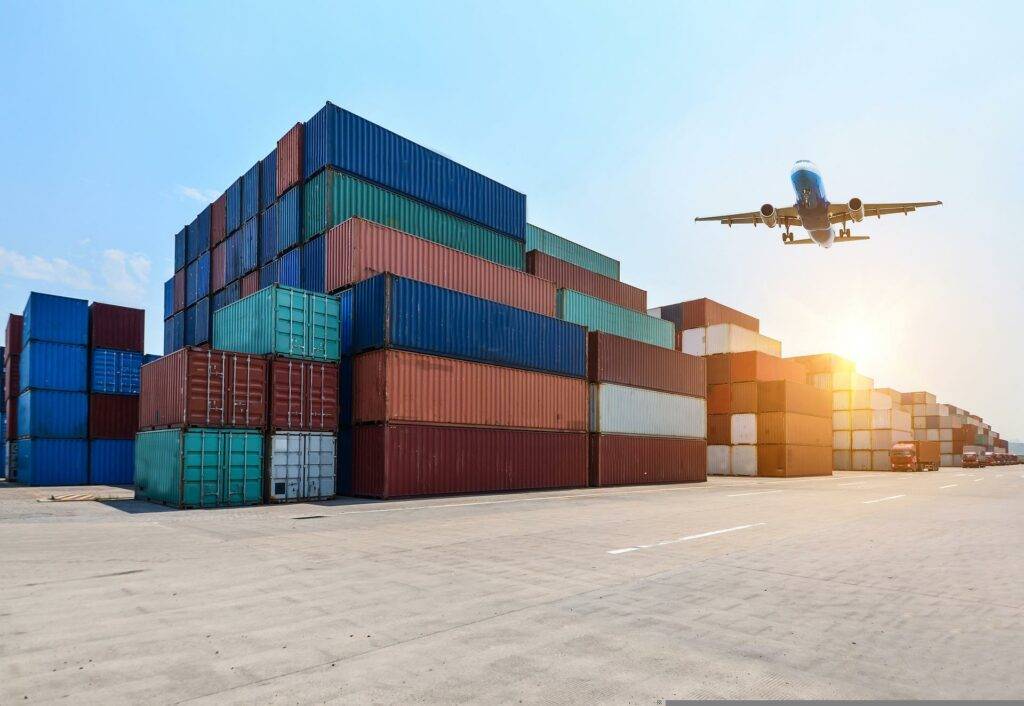 Shipping containers stacked on shore with plane flying overhead